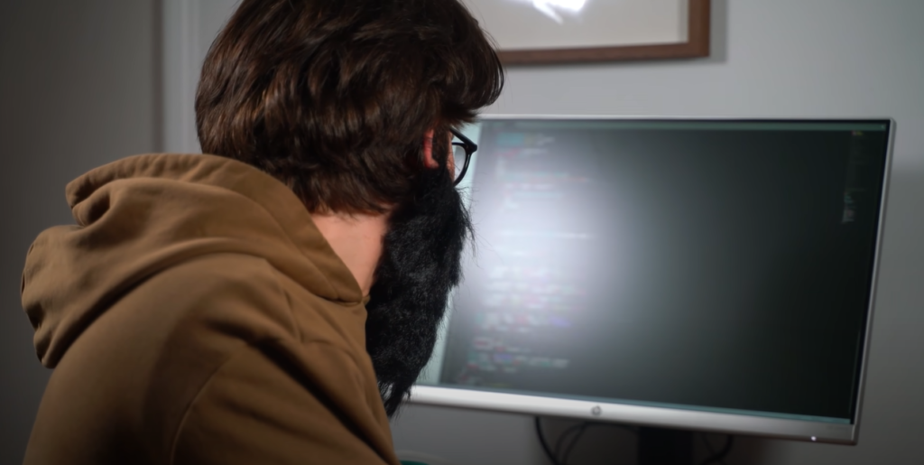 A bearded man sitting in front of a computer screen, with partially blurred lines of code visible.
