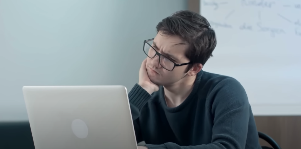 A man wearing eyeglasses in front of a laptop, resting his hand on his chin