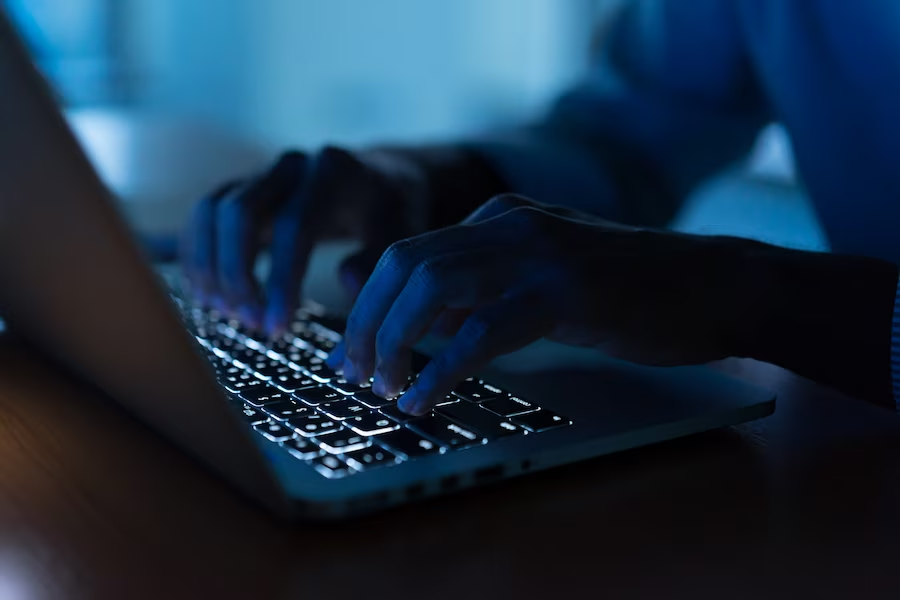 A hand typing on a laptop in a dimly lit room with hints of blue lights